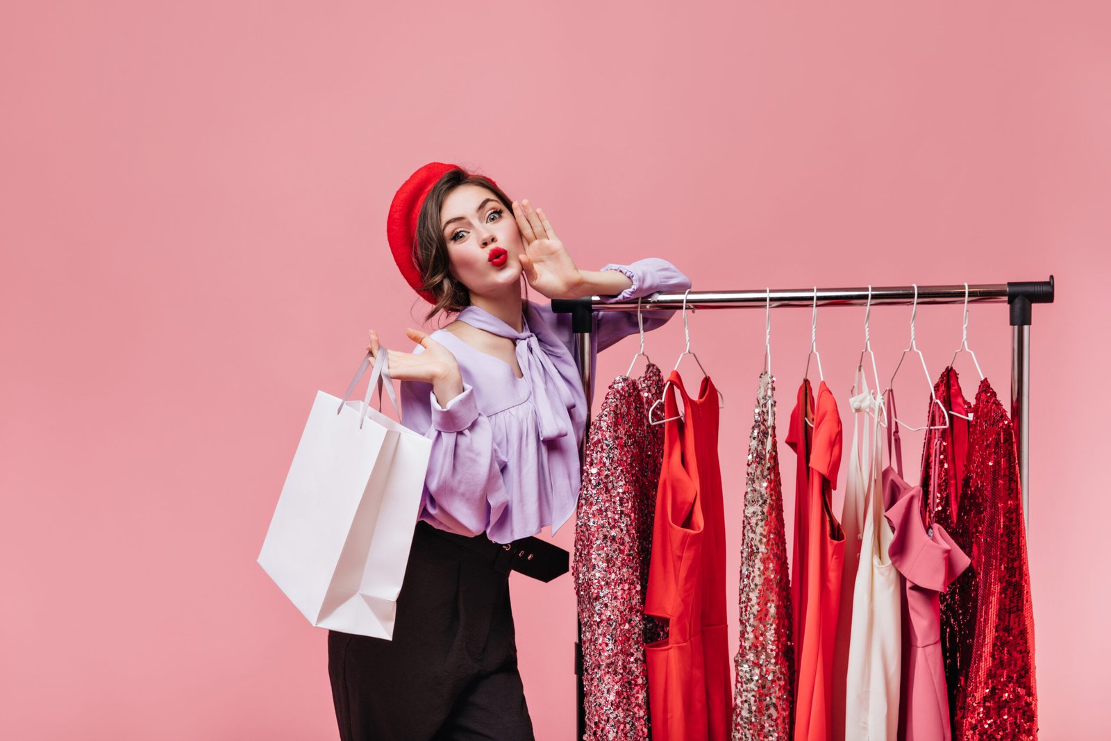 Portrait of girl in red beret whistling and holding package on pink background with hangers with dresses.